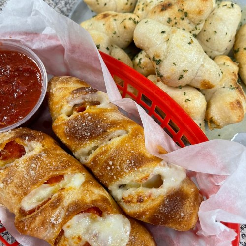A red basket with two cheesy pepperoni rolls and a side of marinara sauce, alongside a tray of garlic knots.