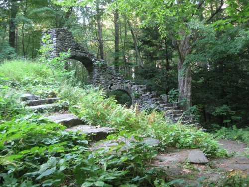Ruins of a stone structure surrounded by lush greenery and trees, with a winding stone path leading through the scene.