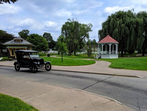 A vintage black car drives past a gazebo in a park with lush greenery and cloudy skies.