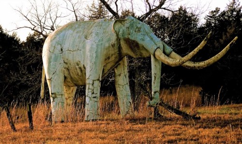 A large, weathered elephant statue stands in a grassy field, surrounded by trees and a sunset backdrop.