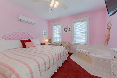 Cozy pink bedroom with a large bed, armchair, and a bathtub, featuring bright windows and decorative accents.
