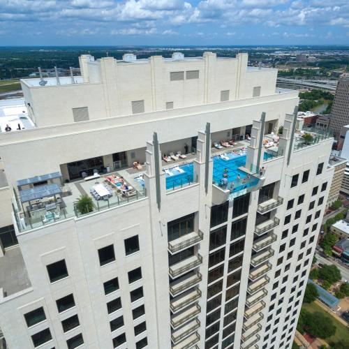 Aerial view of a high-rise building with a rooftop pool and lounge area, surrounded by cityscape and blue skies.