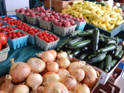 A vibrant market display featuring baskets of tomatoes, potatoes, onions, zucchini, and bananas on a green tablecloth.