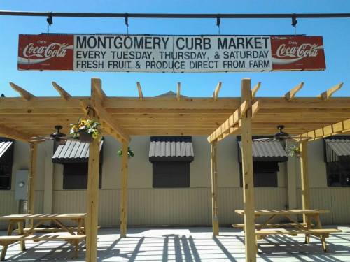 Sign for Montgomery Curb Market, featuring fresh produce, with wooden structures and picnic tables under a clear blue sky.