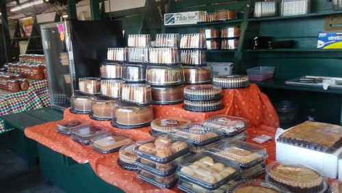 A display of various baked goods in clear containers on a table, with a colorful tablecloth in a market setting.