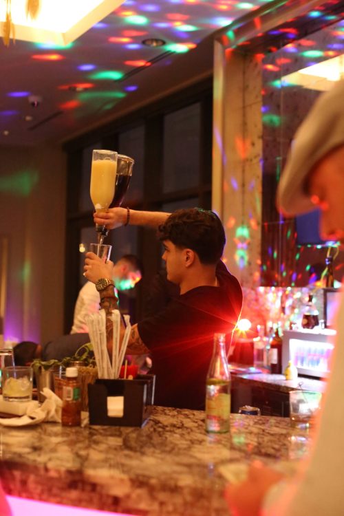 A bartender skillfully pours a large drink at a vibrant bar with colorful lights and patrons in the background.