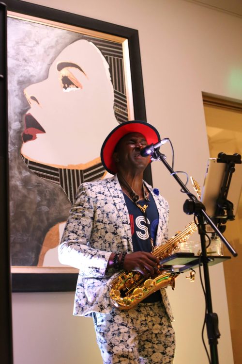 A musician in a patterned suit and red hat plays the saxophone while singing, with a large portrait in the background.