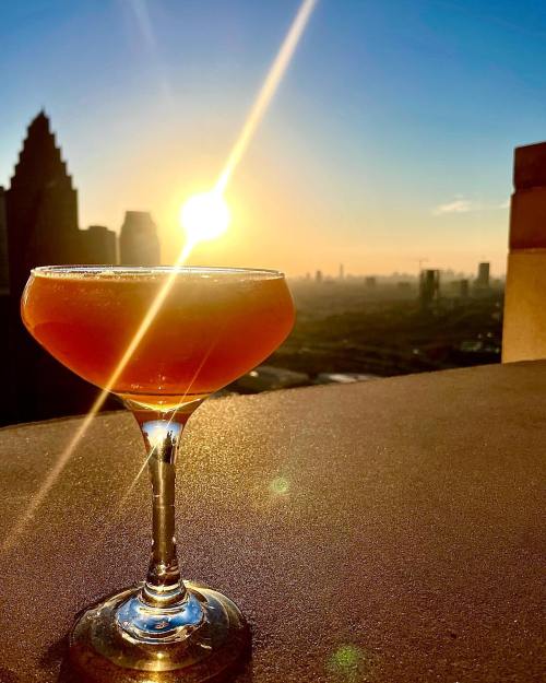 A cocktail glass sits on a ledge with a sunset skyline in the background, casting a warm glow over the scene.