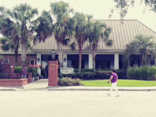 A person in a purple shirt walks past a building surrounded by palm trees and greenery.