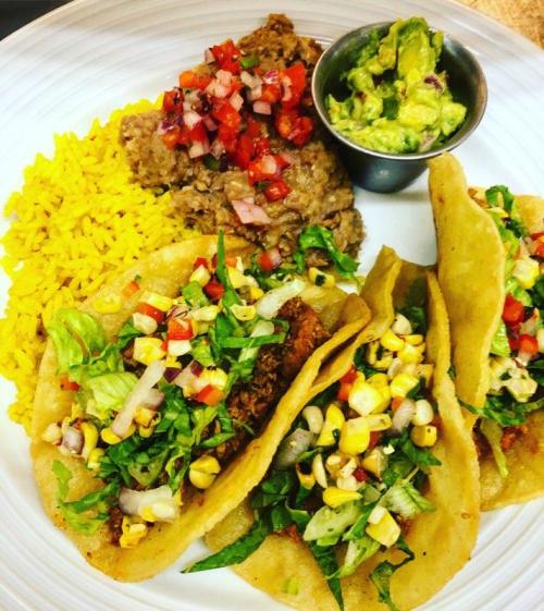 A plate of three tacos filled with lettuce, corn, and salsa, served with yellow rice, refried beans, and guacamole.