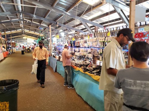 A bustling market scene with vendors selling various items, shoppers browsing, and colorful displays along the aisles.