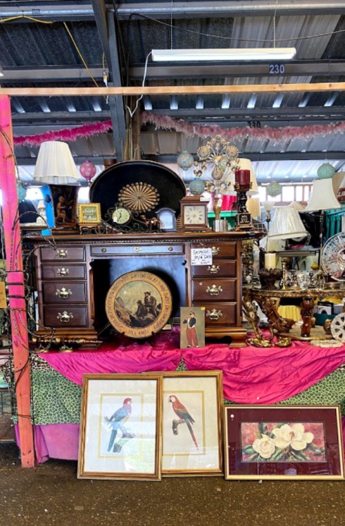 A vintage display featuring a wooden dresser, lamps, clocks, and framed artwork of birds and flowers.