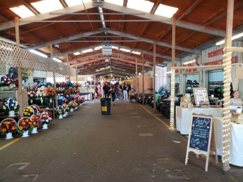 A bustling market interior with stalls displaying flowers, crafts, and decorations, under a wooden roof.