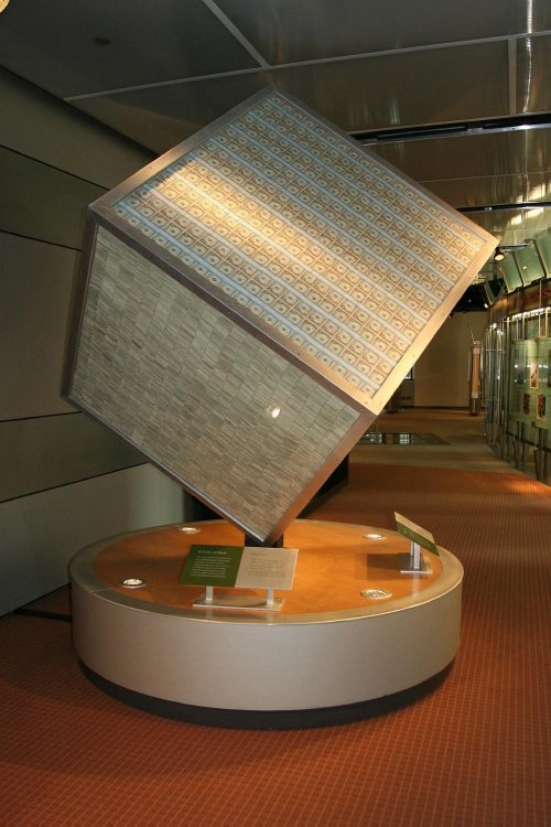 A large, tilted cube sculpture with patterned surfaces, displayed on a circular pedestal in a modern interior space.