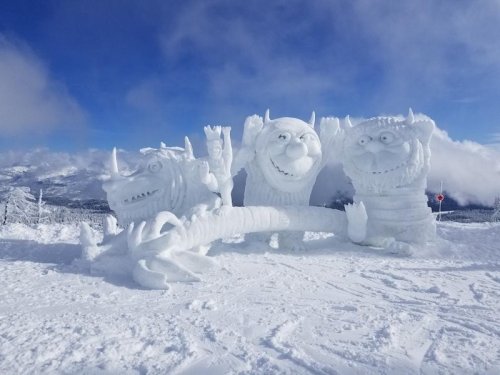 Three large, whimsical snow sculptures resembling friendly monsters, set against a snowy mountain backdrop and blue sky.