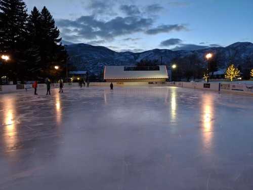 An outdoor ice skating rink at dusk, surrounded by trees and mountains, with skaters enjoying the smooth surface.