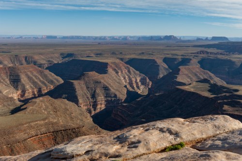 A vast canyon landscape with layered rock formations under a clear blue sky, stretching into the distance.