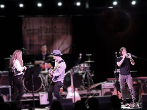 A band performs on stage with a guitarist, a trumpeter, and a vocalist, surrounded by musical instruments and equipment.