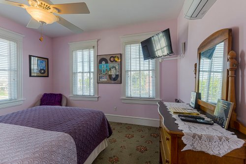 Cozy bedroom with pink walls, a ceiling fan, windows, a TV, and a wooden dresser with decorative items.