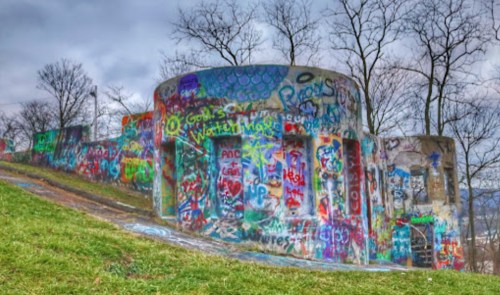 Colorful graffiti covers cylindrical structures on a grassy hill, surrounded by bare trees under a cloudy sky.