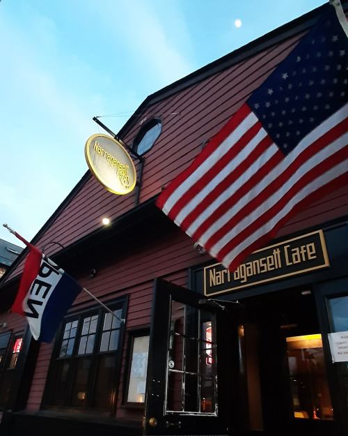 Exterior of Narragansett Cafe with flags, including the American flag, against a twilight sky.