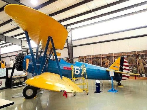 A vintage biplane with a blue body and yellow wings displayed in a hangar, featuring a red and white tail.