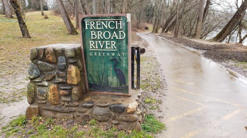 Sign for French Broad River Greenway near a wet, tree-lined path by the river.
