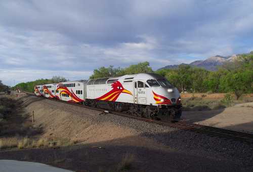 A colorful train with a red and yellow design travels along a track surrounded by greenery and mountains.