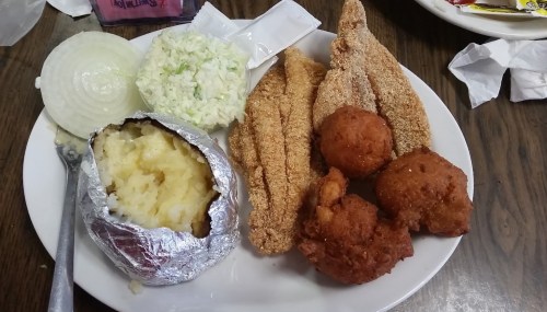 A plate of fried fish fillets, hushpuppies, coleslaw, sliced onions, and a baked potato wrapped in foil.