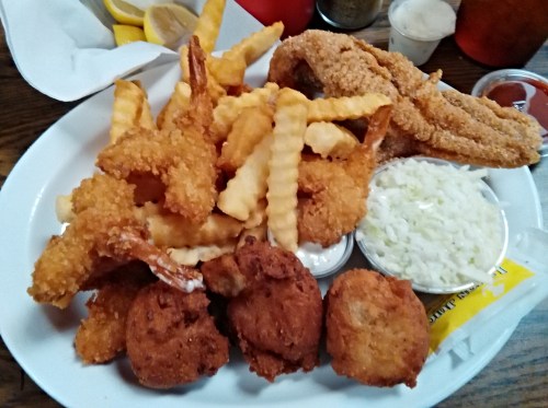 A plate of fried seafood including shrimp, fish, and hushpuppies, served with crinkle-cut fries and coleslaw.