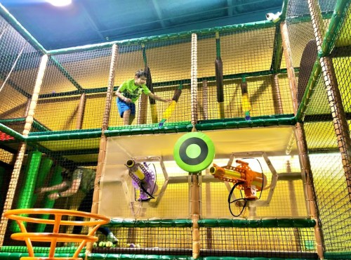 A child plays on a multi-level indoor playground, navigating nets and aiming at targets with toy blasters.
