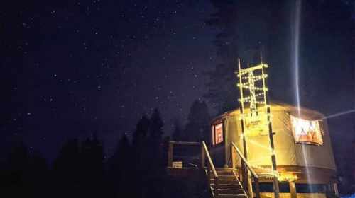 A cozy, illuminated tiny house under a starry night sky, surrounded by trees.