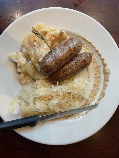 A plate with two sausages, sauerkraut, and a side of creamy potato salad, garnished with a bay leaf.