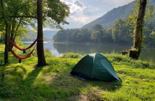 A green tent and two hammocks are set up by a serene river, surrounded by trees and mountains under a sunny sky.