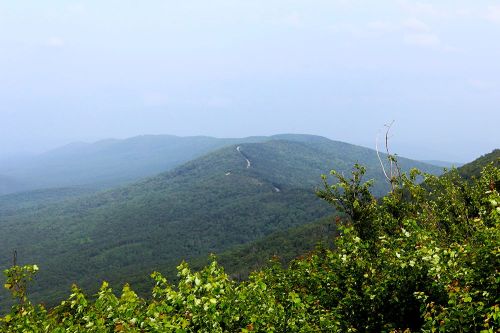 A panoramic view of rolling green mountains under a hazy sky, with a winding road visible in the distance.