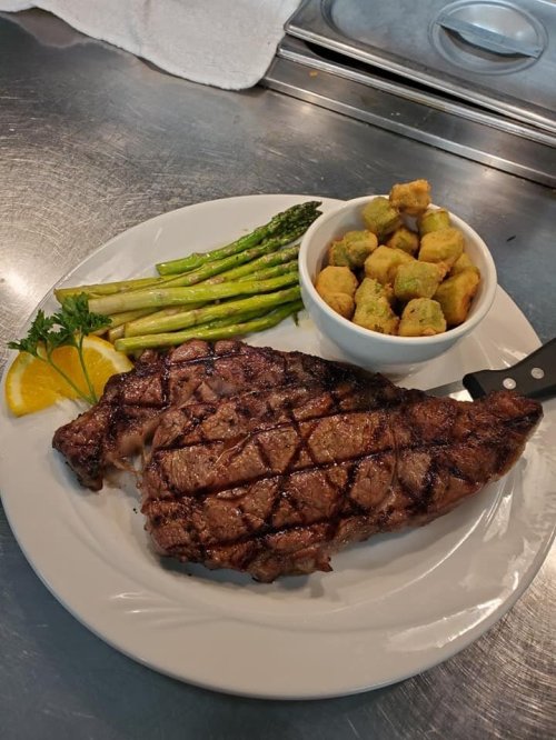 A grilled steak with a crisscross pattern, served with asparagus, fried okra, and lemon wedges on a white plate.