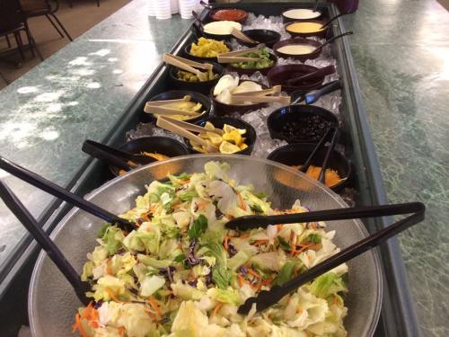 A salad bar featuring a large bowl of mixed greens and various toppings in bowls on ice, with serving utensils.