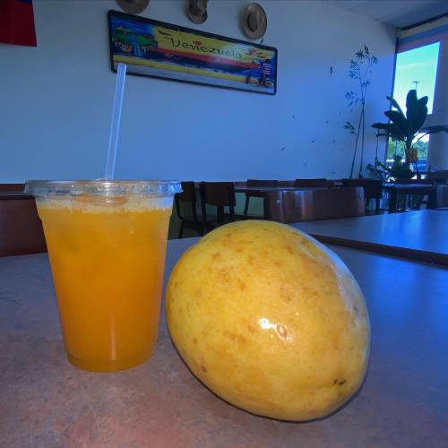 A cup of orange juice next to a large, ripe mango on a table in a brightly lit room.
