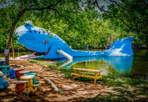 A large blue whale sculpture by a pond, featuring a slide and surrounded by trees and colorful seating.