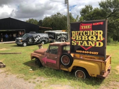 An old truck with a sign for "The Butcher BBQ Stand" parked outside a building, with Jack Daniel's vehicles nearby.