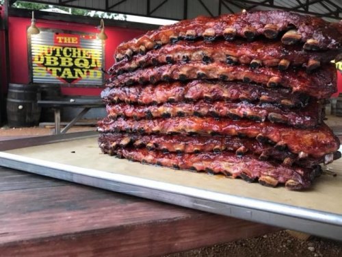 A towering stack of glazed ribs on a tray, with a colorful sign for "The Butcher Ribbon Stand" in the background.