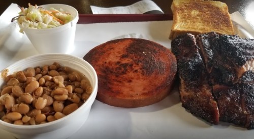 A plate with grilled meat, a slice of burnt bologna, baked beans, coleslaw, and toasted bread on a tray.