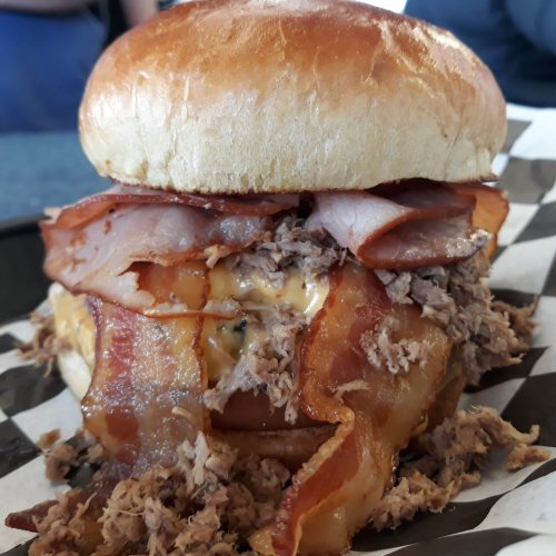 A stacked burger with pulled pork, bacon, and melted cheese, served in a soft bun on a checkered paper plate.