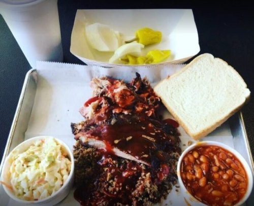 A tray with barbecue meat, sauce, coleslaw, baked beans, pickles, and a slice of bread, alongside a cup.