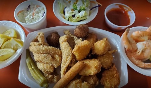 A platter of fried seafood, including fish, shrimp, and hushpuppies, with sides of coleslaw, salad, and lemon wedges.