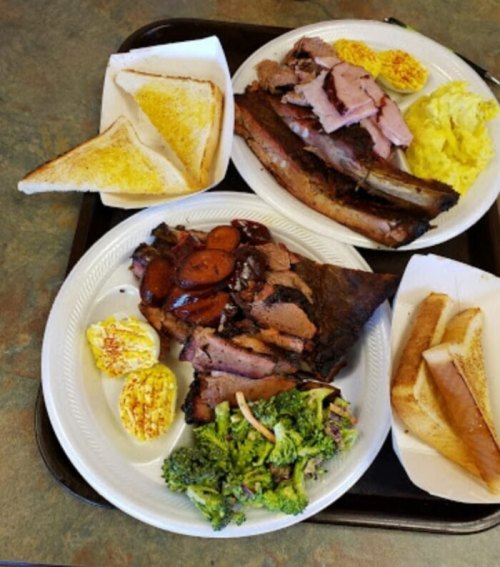Two plates of barbecue featuring sliced meats, toast, scrambled eggs, and broccoli on a tray.