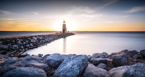 A lighthouse stands on a rocky pier at sunset, with calm waters reflecting the warm colors of the sky.