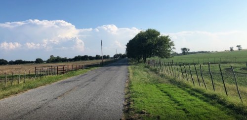 A quiet rural road lined with fences, fields, and trees under a clear blue sky with fluffy clouds in the distance.