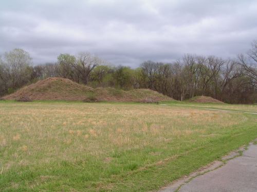Two grassy mounds in a field, surrounded by trees under a cloudy sky. A path runs alongside the mounds.