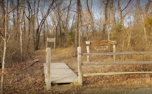 A wooden bridge leads into a wooded area with a sign marking the entrance to a nature trail.
